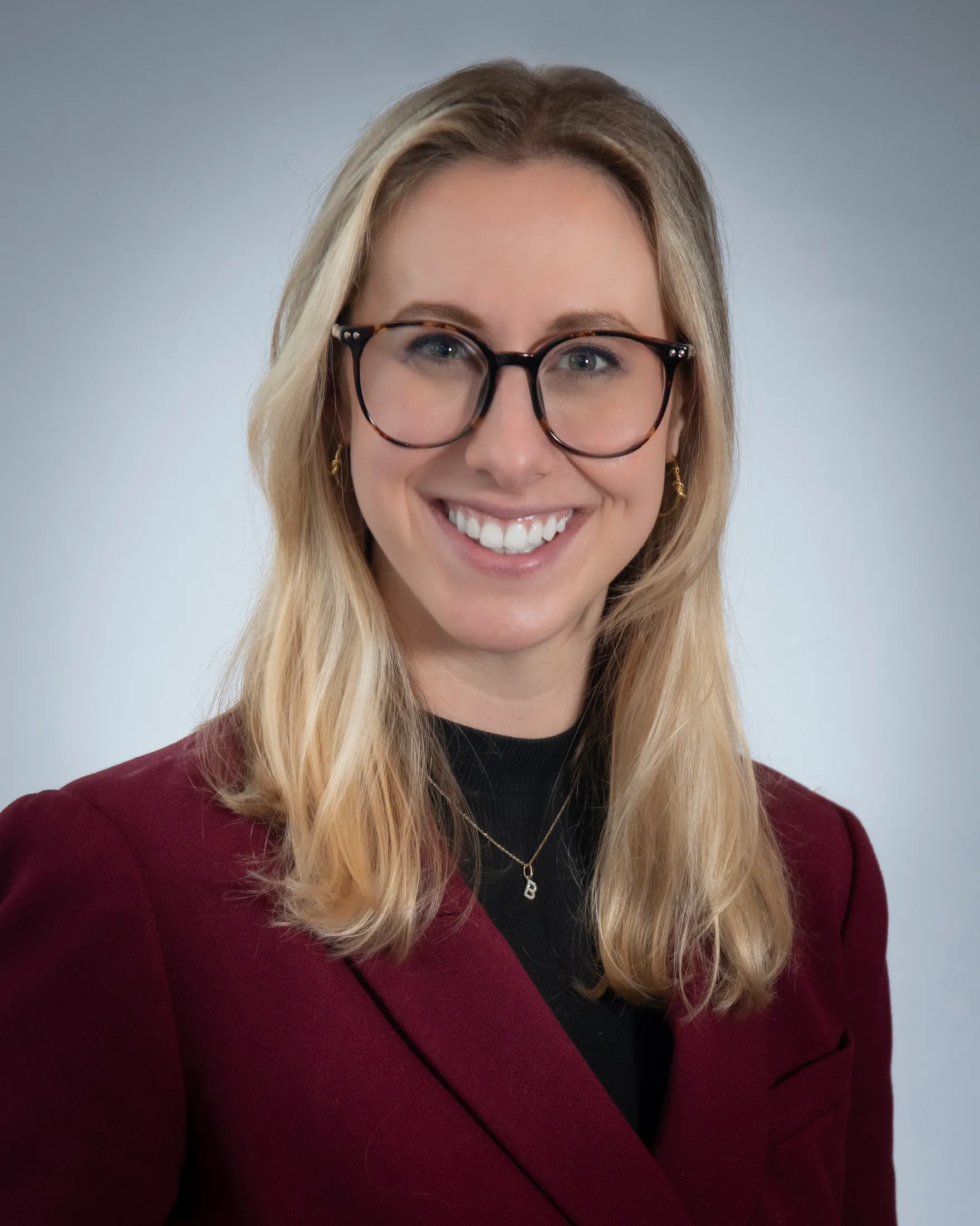 A smiling woman wearing dark-framed glasses and a burgundy jacket against a gray background.