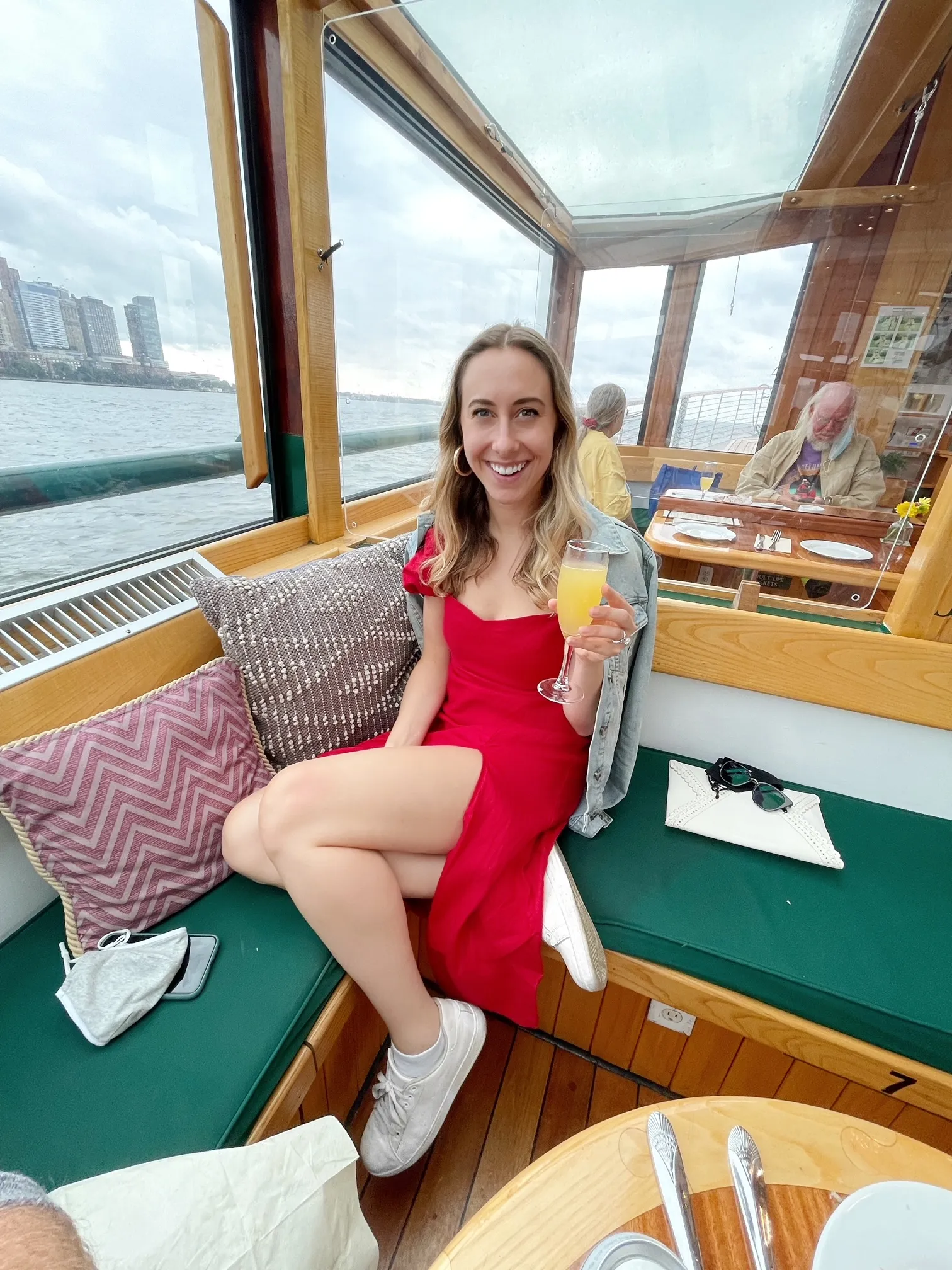 A woman in a red dress holding a glass aboard a boat with water and a city skyline outside.