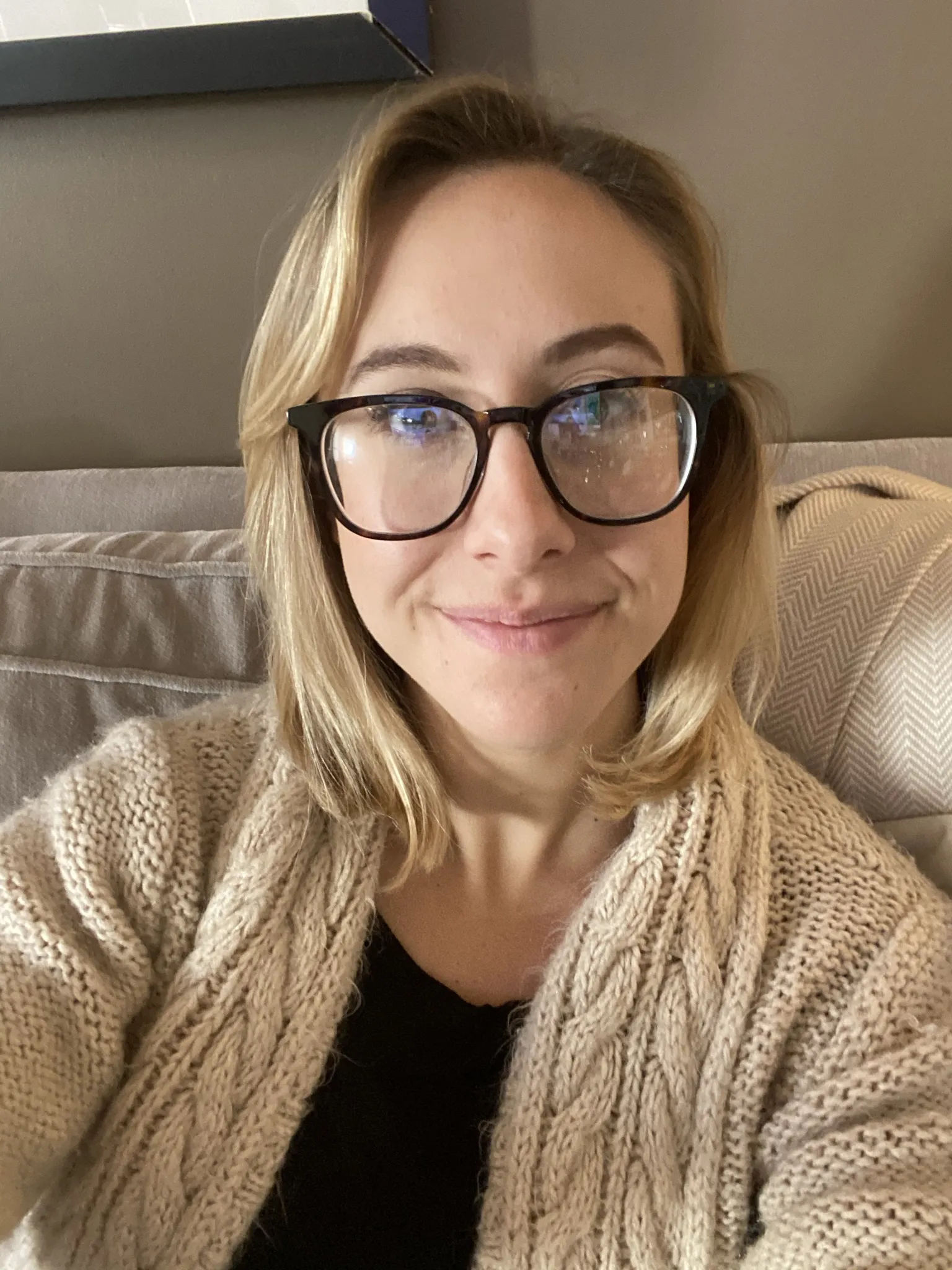 A woman in glasses and a knitted cardigan smiling from a sofa.