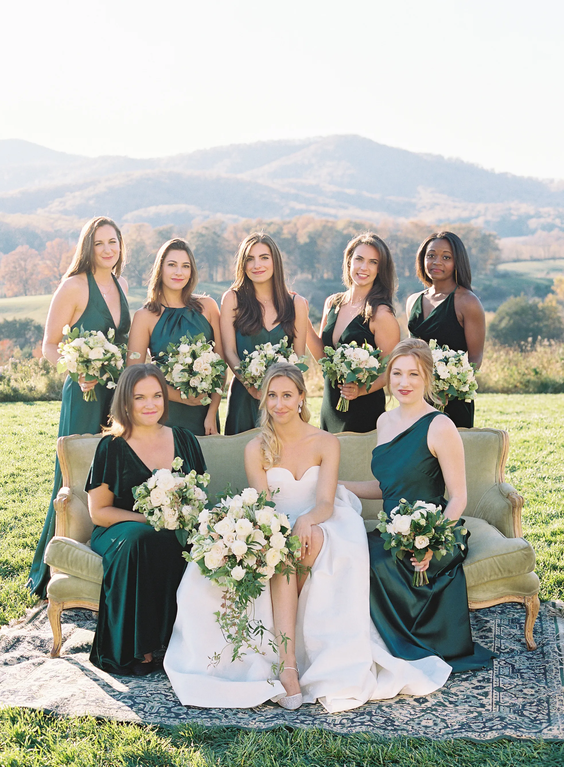 A bride and a group in green dresses posing outdoors with hills in the distance.