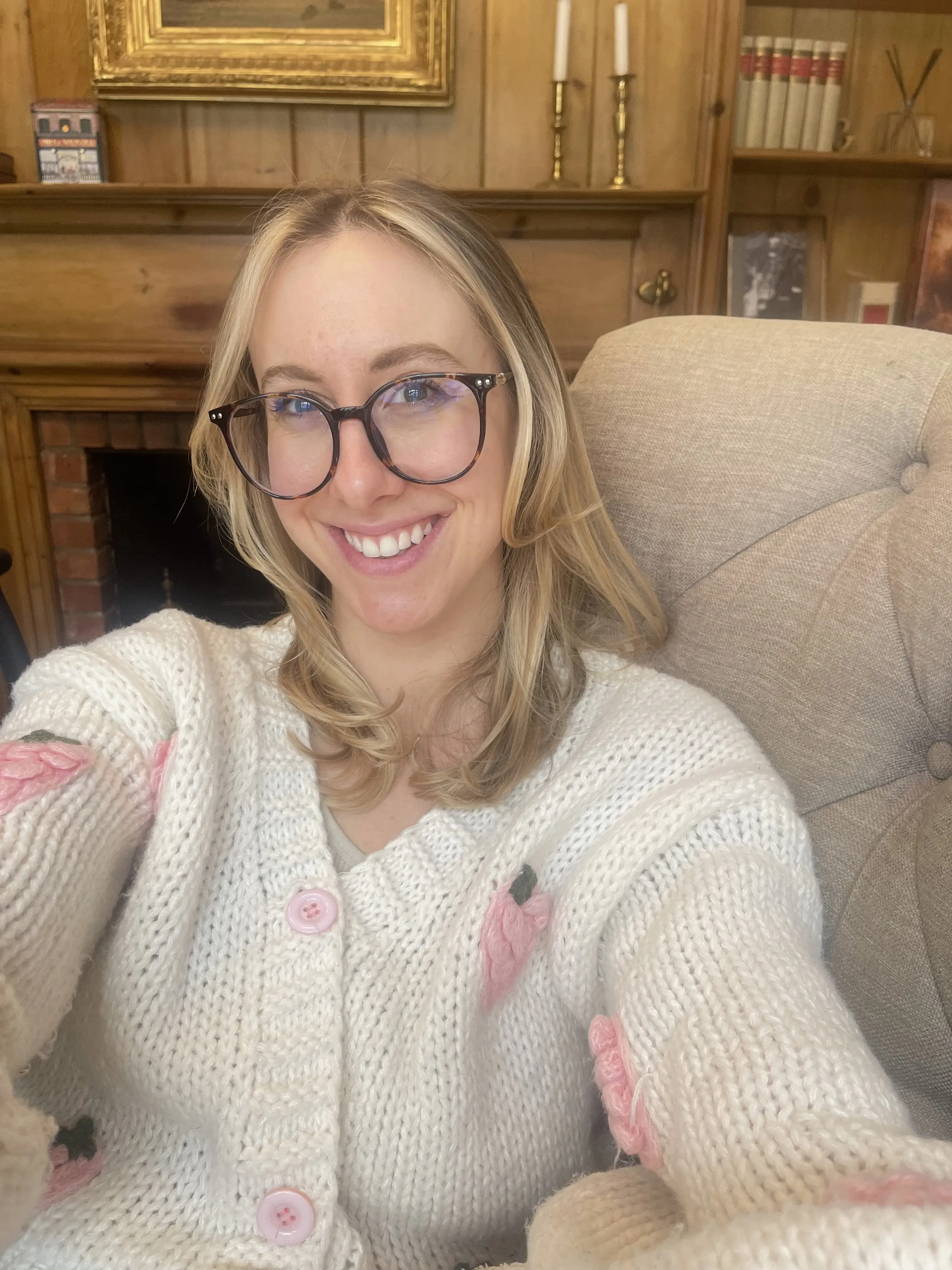 A smiling woman in glasses and a pale cardigan beside a fireplace.