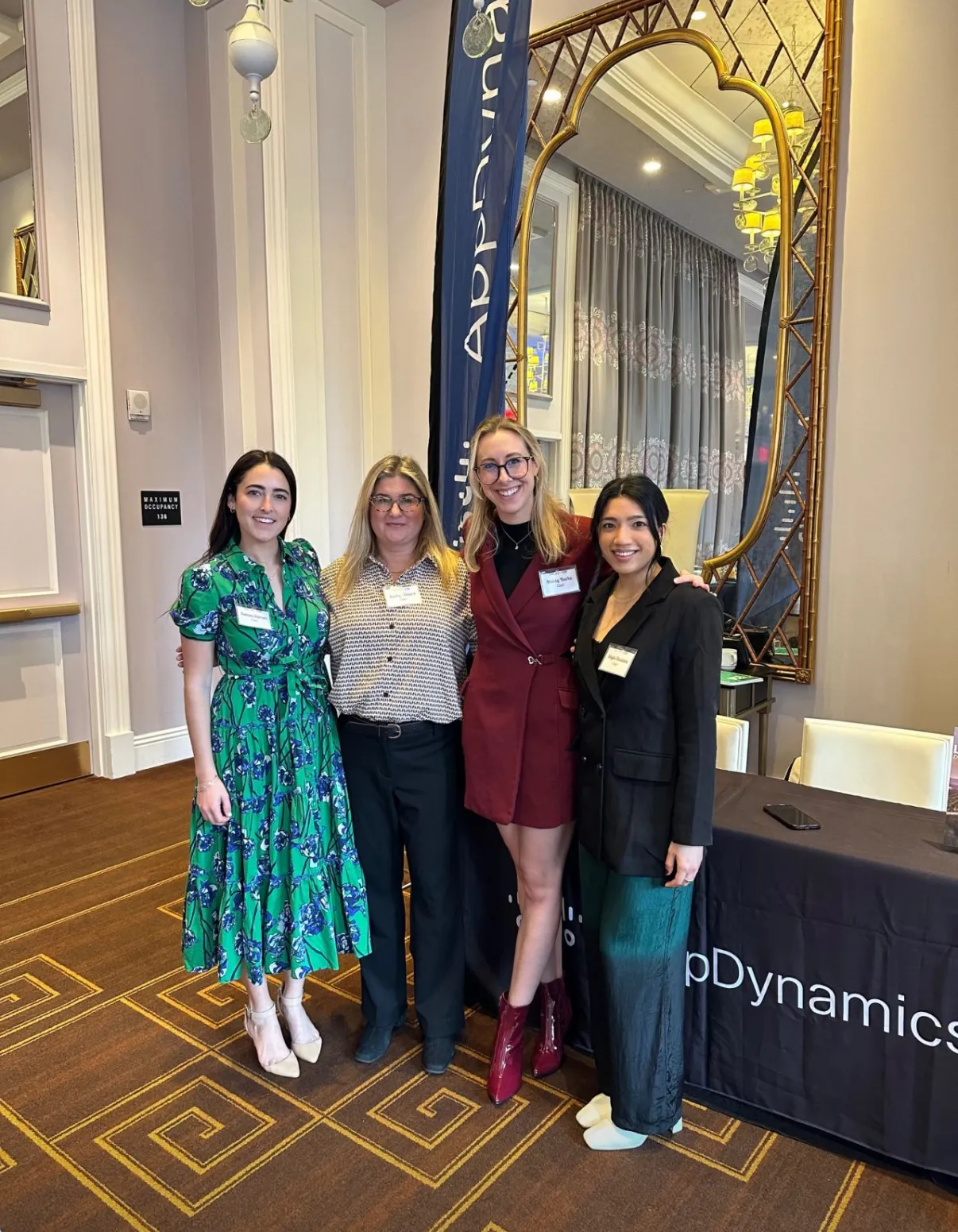 Four women posing in a grand room beside a large gold-framed mirror.