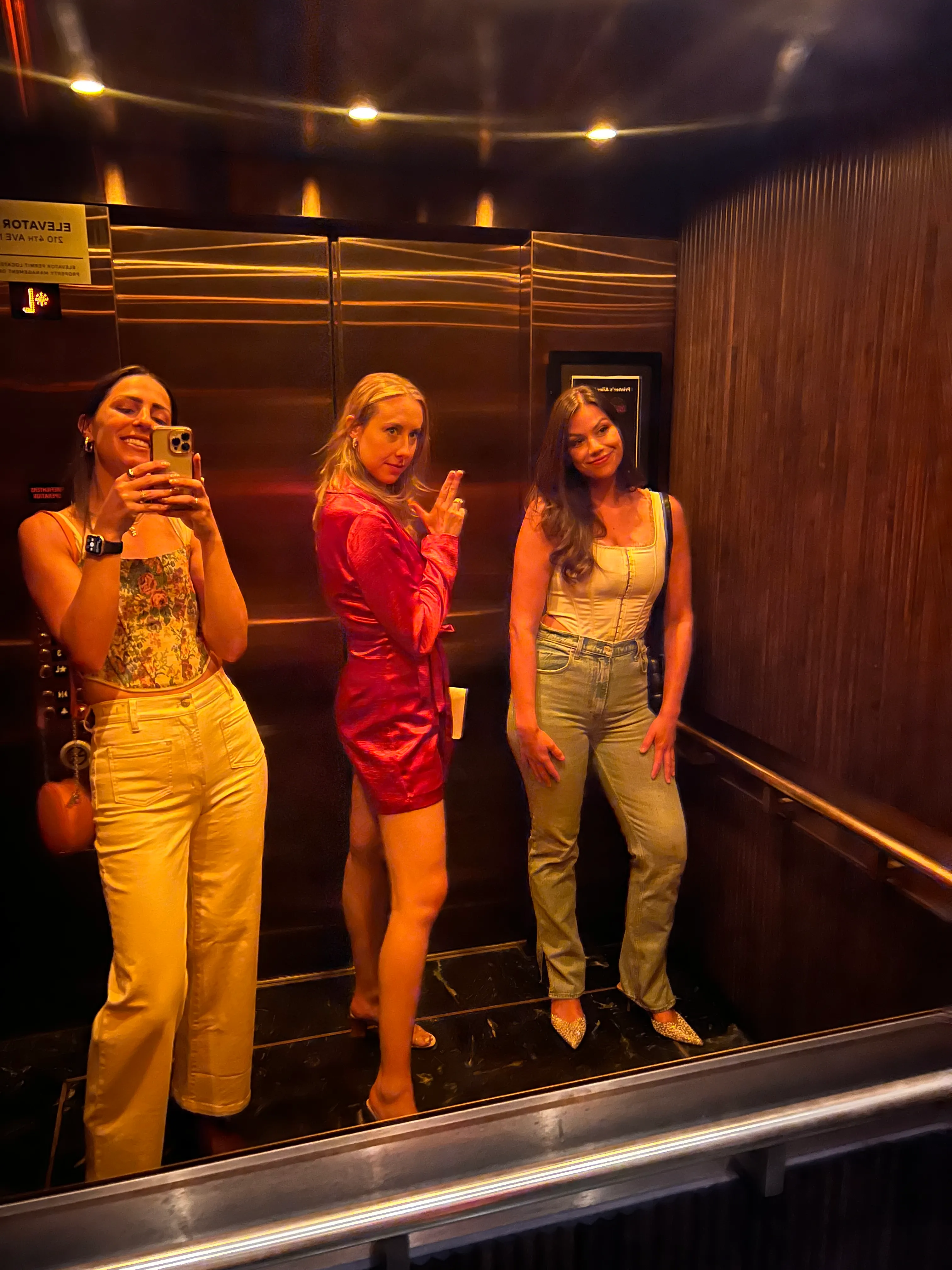 Three women posing for a mirror photograph in a warmly lit elevator.