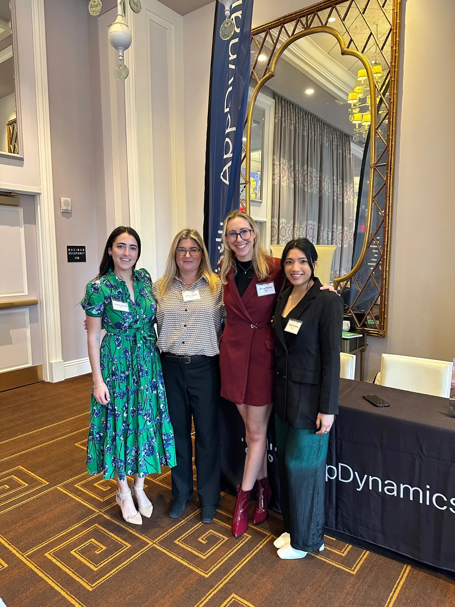 Four women posing together beneath a tall gold-framed mirror at an event.