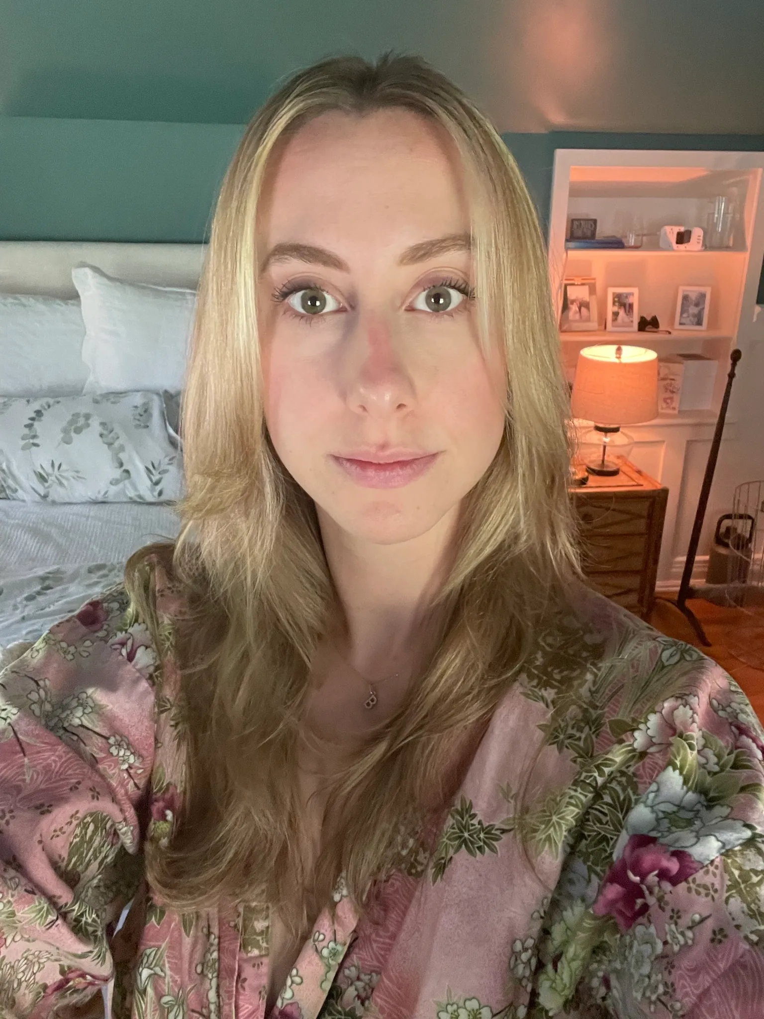 A woman in a floral robe taking a portrait in a softly lit bedroom.