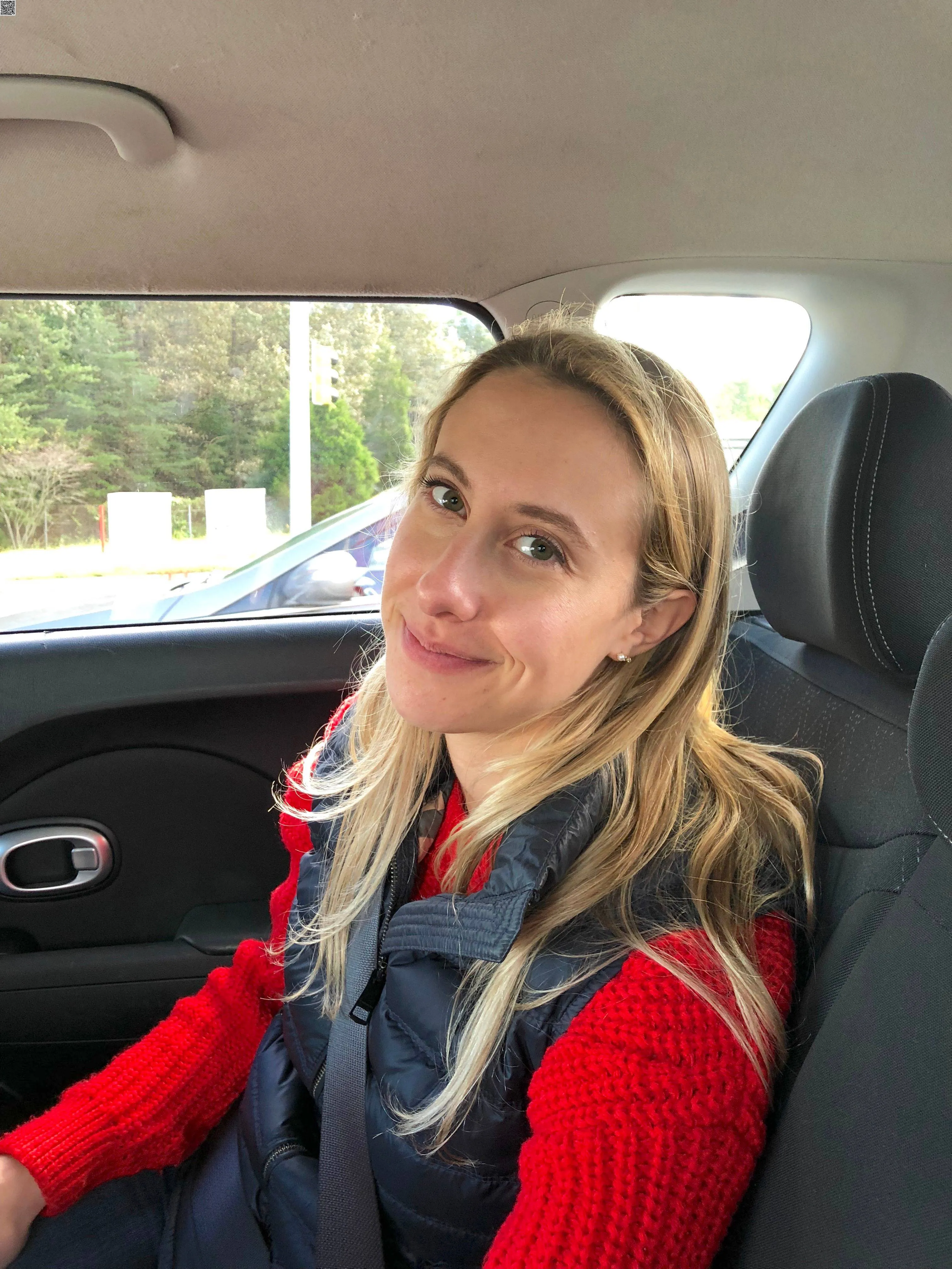 A woman in a red sweater and dark vest sitting in a car.