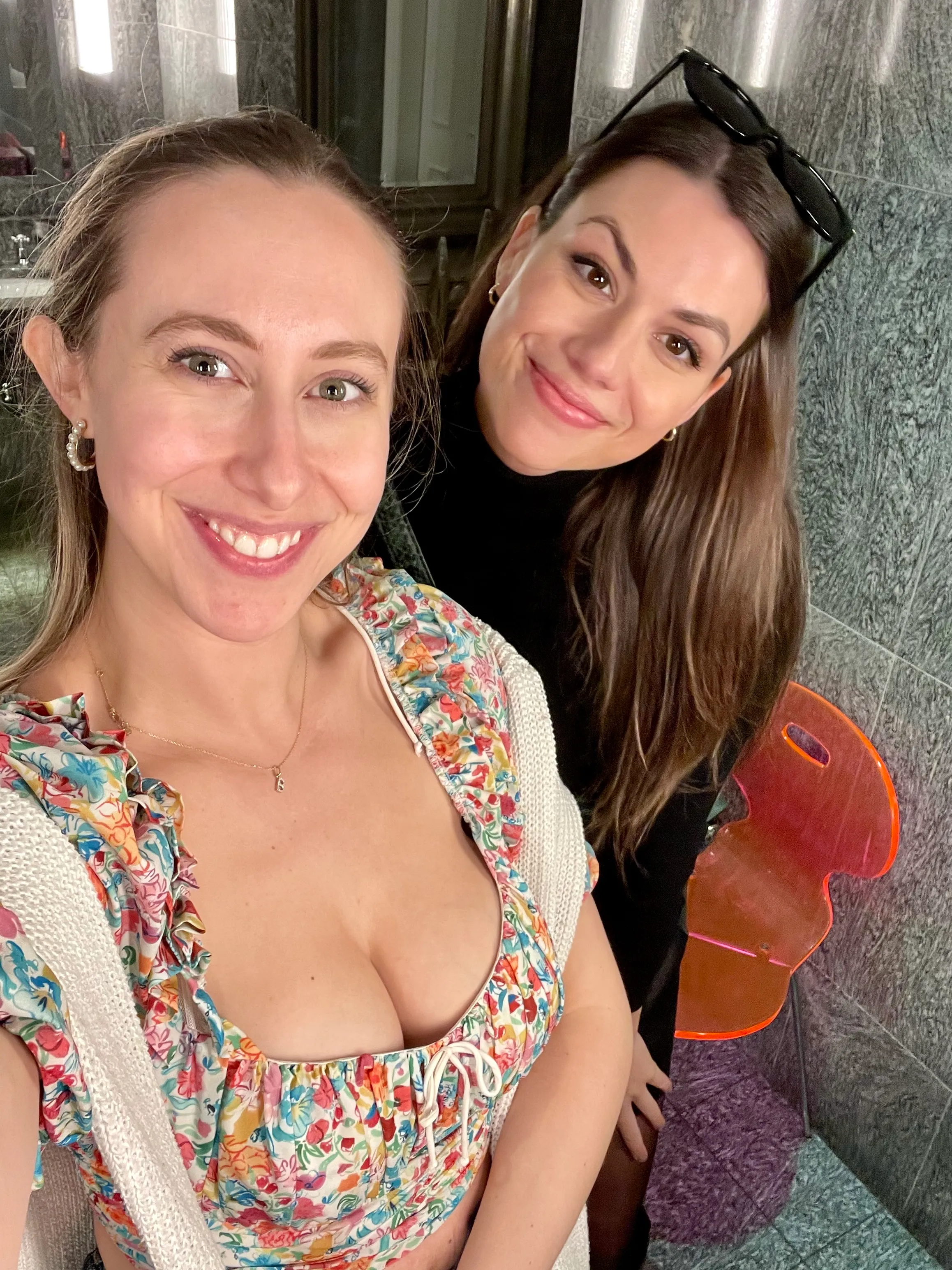 Two women smiling together indoors, one wearing a colorful floral outfit.