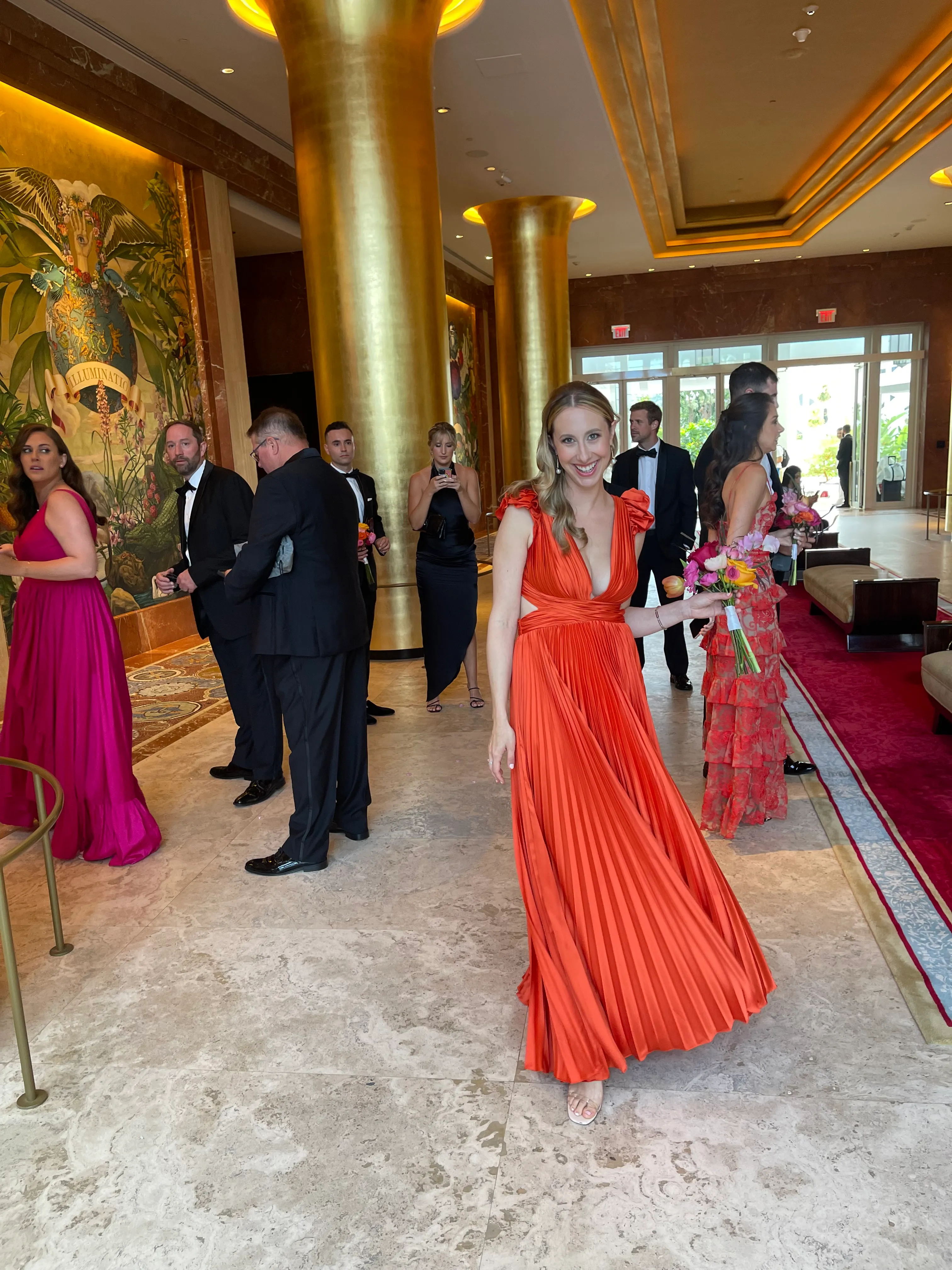 A smiling woman in an orange gown holding flowers beneath golden columns.