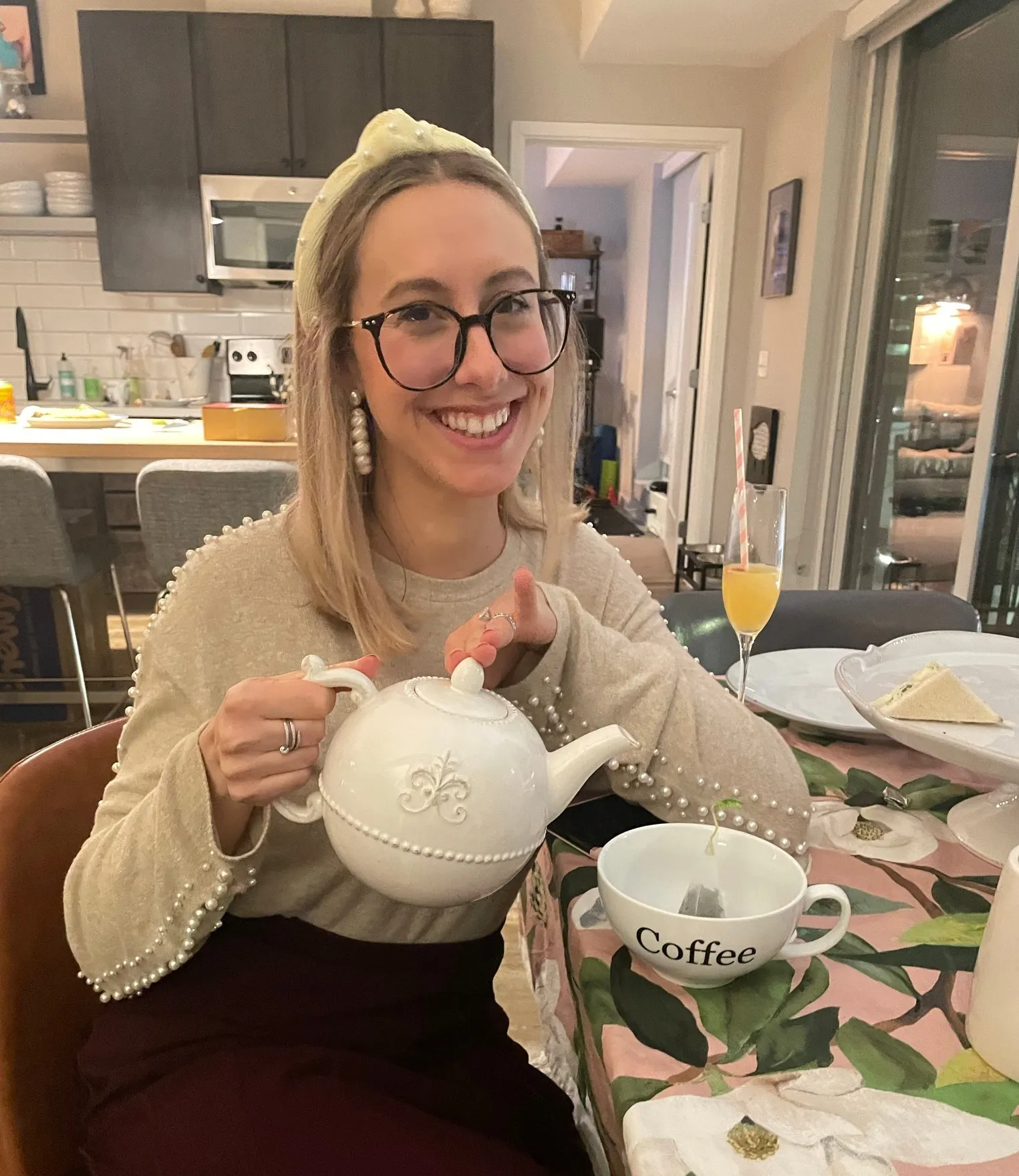 A smiling woman holding a white teapot at a table set for afternoon tea.