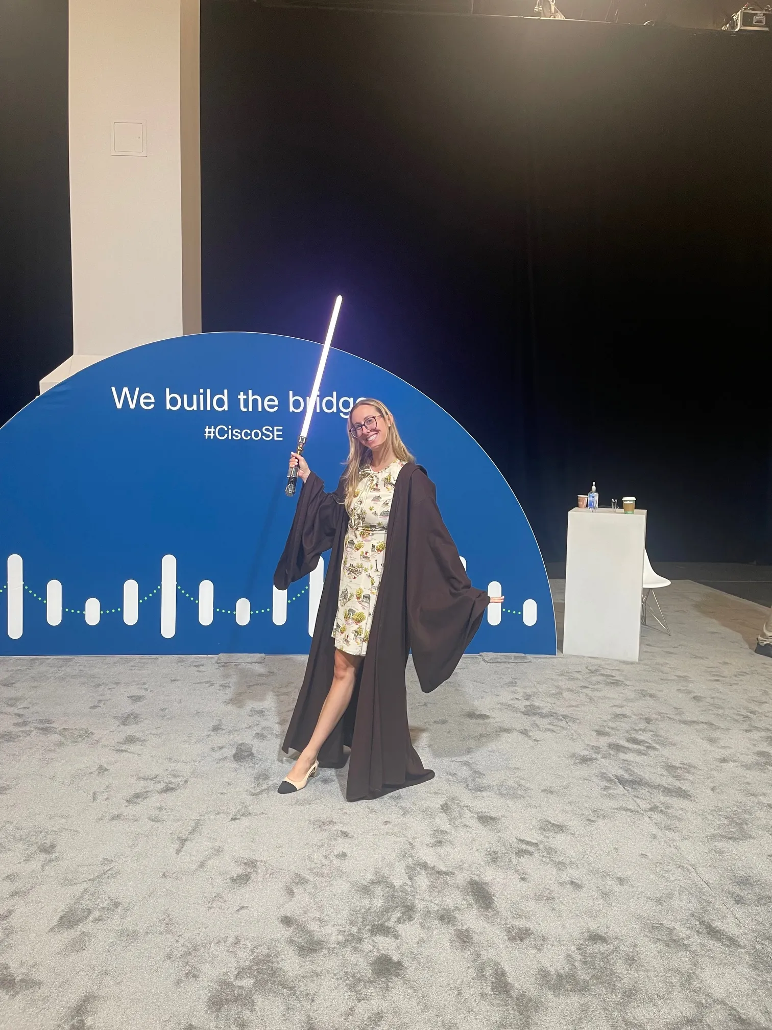 A smiling woman wearing a dark robe and holding a glowing lightsaber at an event.