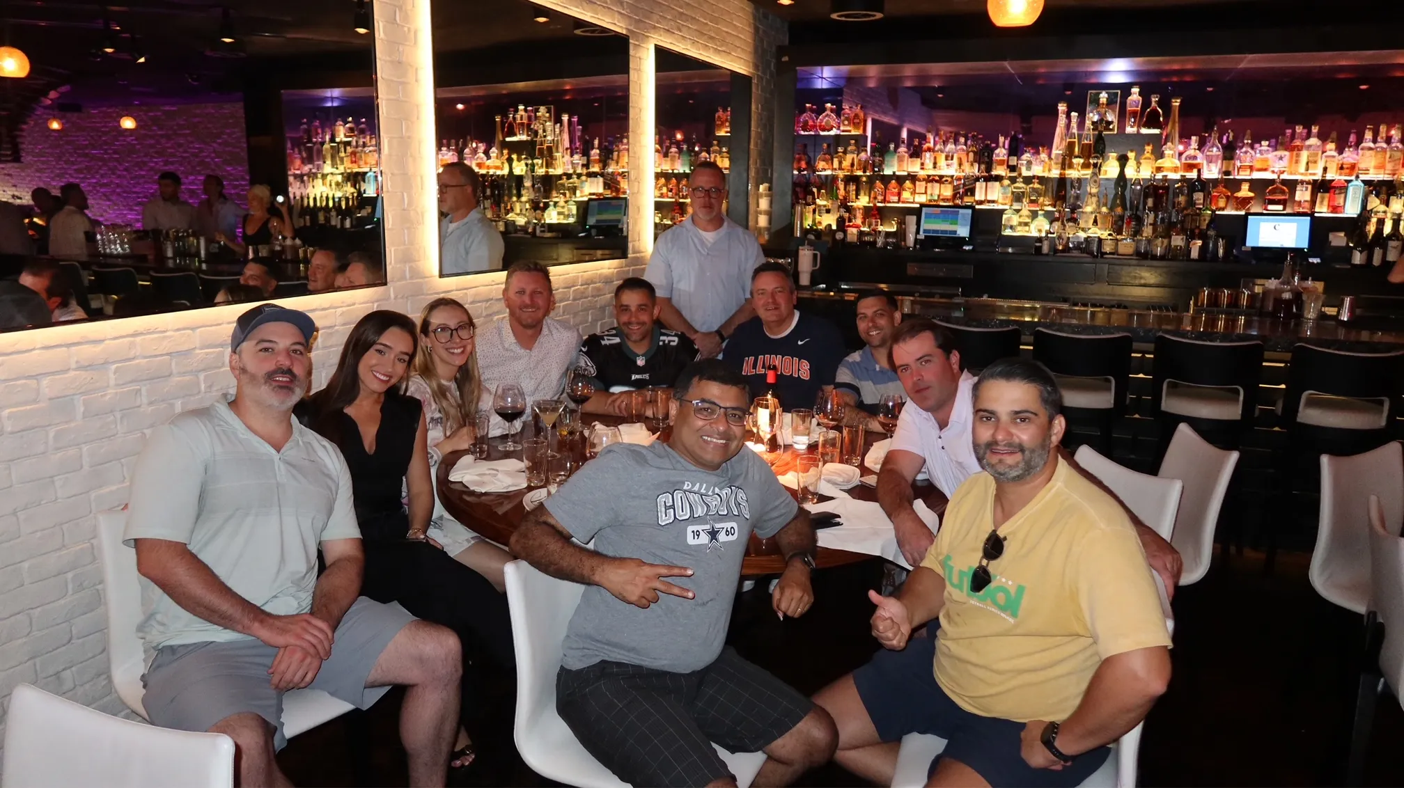 A large group gathered around a restaurant table with a brightly lit bar behind them.
