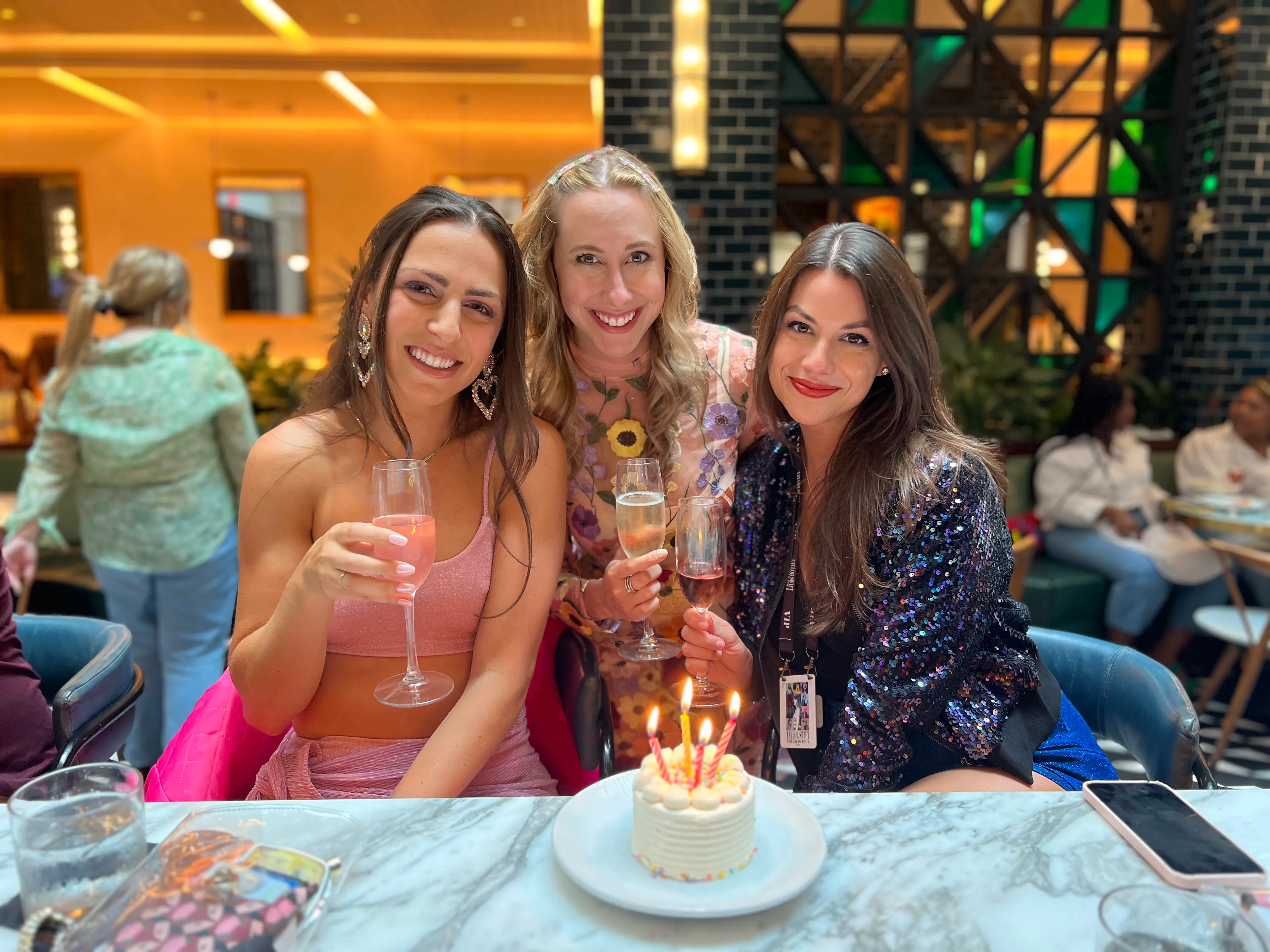 Three women raising glasses beside a small cake with lit candles.